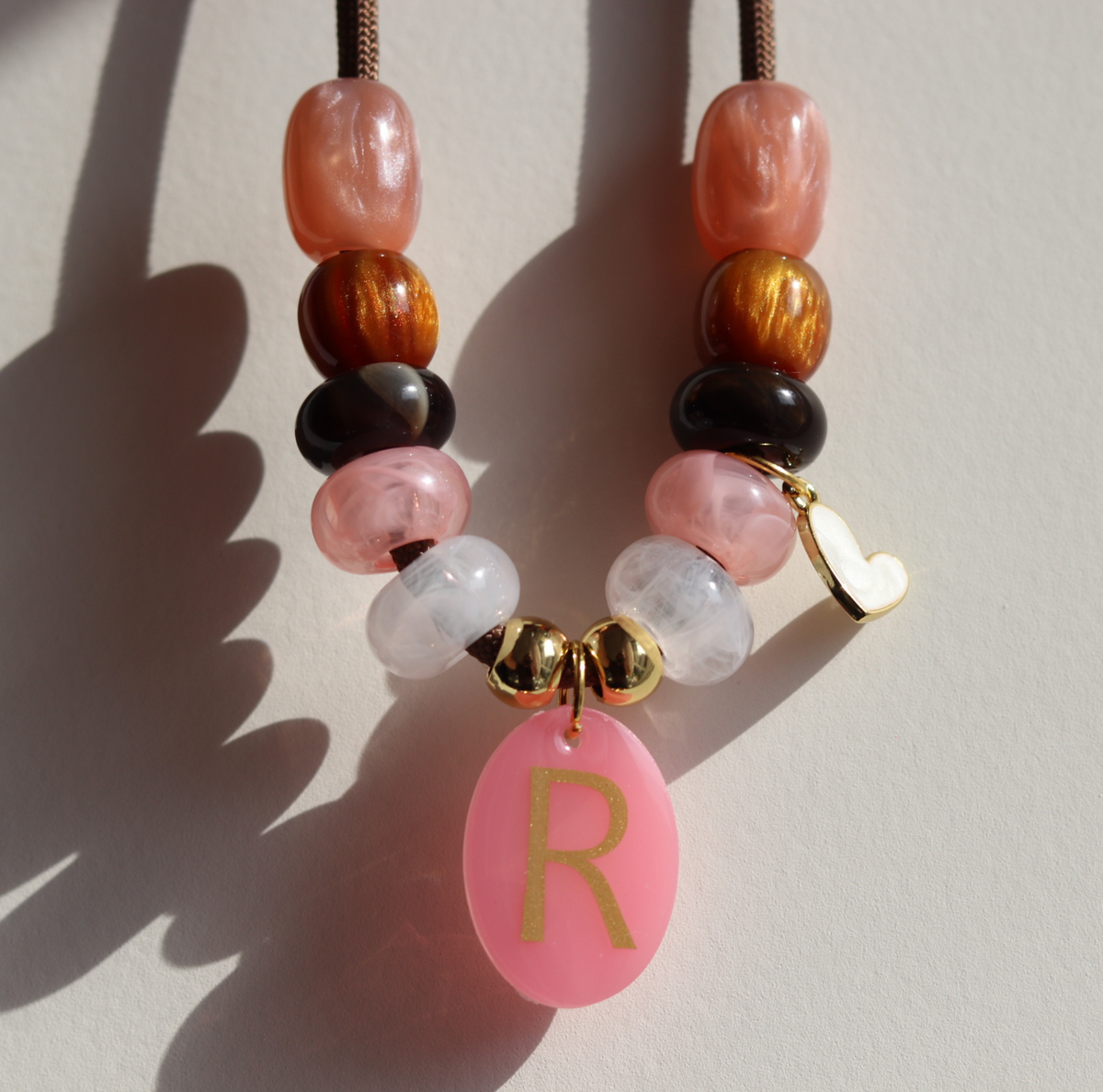 Deze handgemaakte hondenketting 'Rosé glow' is opgebouwd uit een stevig bruin koord met een stijlvolle mix van kralen. Het ontwerp combineert glanzende warmroze, zachtroze en marmerwitte kralen met diepbruine en goudkleurige accenten voor een luxe uitstraling. en een penning met het initiaal van je hond