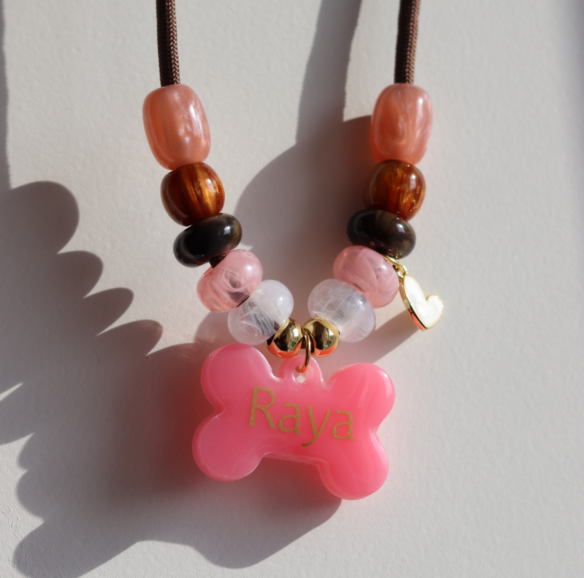 Handgemaakte hondenketting 'Rosé glow' van bruin koord met roze, witte en bruine kralen, een goud hartje en een gepersonaliseerde roze botpenning.