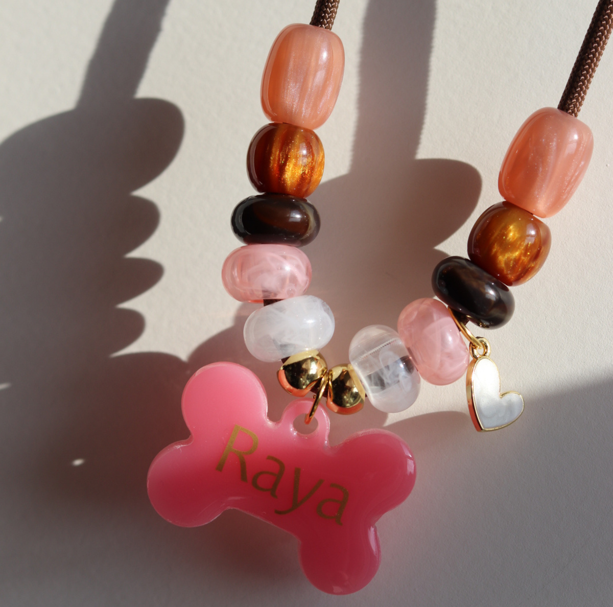 Gepersonaliseerde hondenketting met glanzende rosé, marmerwitte en gouden kralen aan een bruin koord, inclusief goud hartbedeltje en opvallende roze naam-penning in de vorm van een botje.