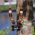 Een handgemaakte, kleurrijke hondenhalsketting met kralen, hangend aan een boomstam in de buitenlucht met een wazige achtergrond van water en groen. De ketting is gemaakt van een bruin-wit gemêleerd koord en is versierd met symmetrisch geregen kralen in de kleuren roestoranje, donkerbruin, crème, glanzend amber, groen en okergeel. Aan de rechterkant hangt een klein, goudkleurig sterretje. Onderaan hangt een donkerbruine hanger in de vorm van een hondenbotje met de gepersonaliseerde naam "Jack" in witte lett