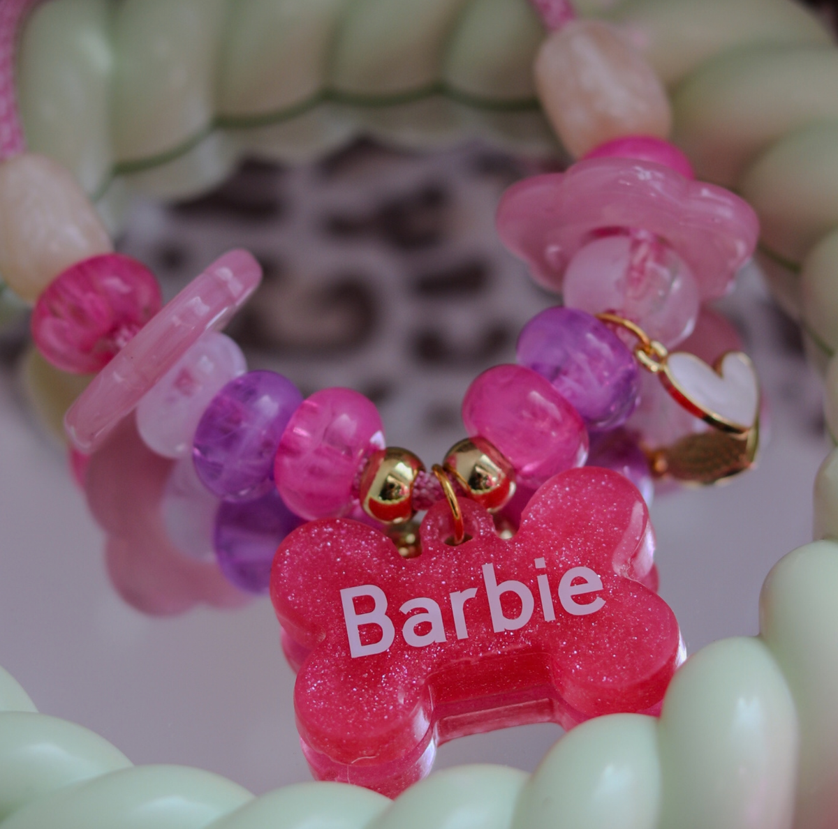 Een kleurrijke en glinsterende hondenhalsketting met kralen, gefotografeerd op een ronde spiegel die omringd is door een gedraaide, lichtgroene rand, met een luipaardprint op de achtergrond. De ketting heeft een roze-wit gemêleerd koord en is versierd met kralen in verschillende tinten roze en paars, waaronder bloemvormige kralen, evenals lichte parelmoerkralen en een goud-wit hartjesbedeltje. Onderaan de ketting, bevestigd met kleine goudkleurige kraaltjes en ringetjes, hangt een glitterroze hanger in de v