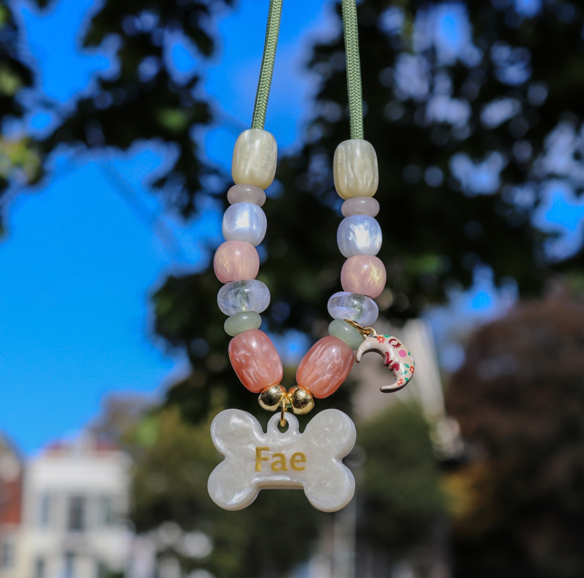 Een hondenkSubtiel hondenkettingje aan een saliegroen koord, geregen met pastelkleurige parelmoerkralen in roze, lila, wit en zalmroze. Onderaan hangt een parelmoerwit hondenbotje met de opgedrukte gouden naam "Fae", twee gouden bolletjes en een klein halvemaantje met bloemmotief.